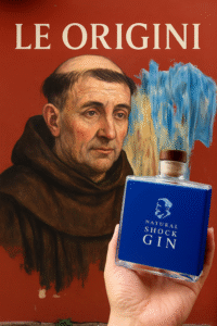 Bottiglia di Tesla Gin davanti a un murale con un frate medievale e la scritta “Le Origini”, richiamo alle radici storiche del gin intorno all’anno Mille.
